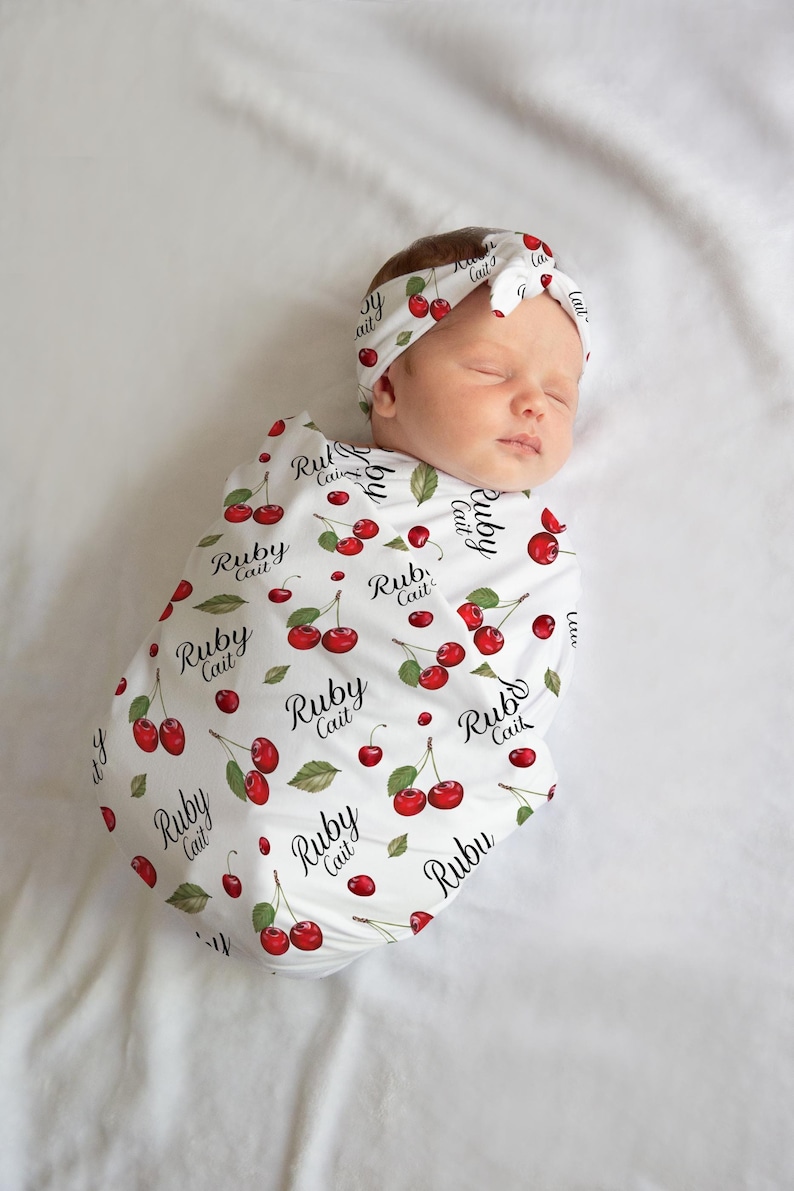 K&ouml;nnte beinhalten: Eine wei&szlig;e Swaddle-Decke mit einem roten Kirschmuster und dem Text "Ruby Cait", der sich wiederholt. Ein Baby ist in die Decke gewickelt und tr&auml;gt ein passendes Stirnband.