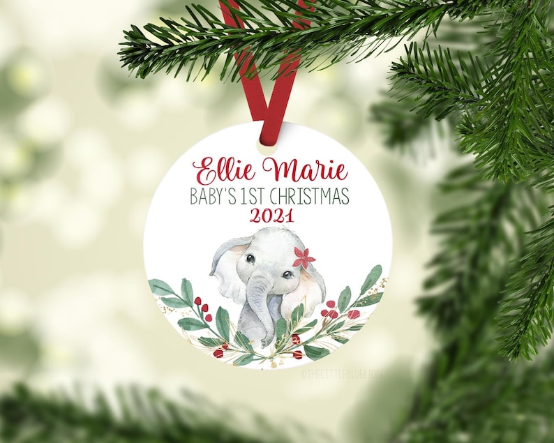 May include: A white Christmas ornament with a red ribbon, featuring the text "Ellie Marie Baby's 1st Christmas 2021." The ornament has a watercolor illustration of a gray elephant with a red flower and green foliage.