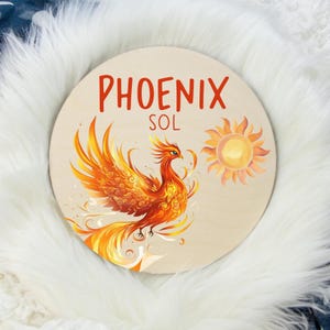 May include: Round wooden sign featuring a vibrant phoenix with orange, yellow, and red feathers, and a watercolor sun. The sign has the words "PHOENIX SOL" in red lettering. The background is a light wood color.