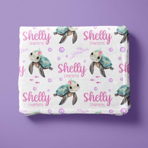 May include: White baby blanket with a repeating pattern of pink and green sea turtles with flowers on their heads. The blanket has the name "Shelly Charlotte" repeated throughout.