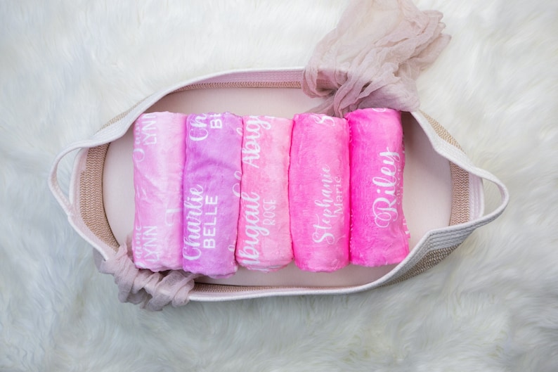 May include: A wicker basket filled with six pink and white ombre baby blankets, each with a different name embroidered on them:  'Lynn', 'Charlie Belle', 'Abigail Rose', 'Stephanie Marie', and 'Riley'.