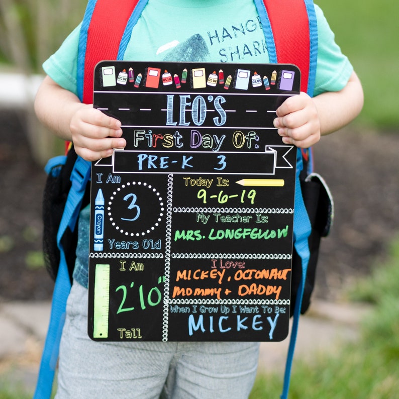 May include: A black chalkboard sign with colorful accents, announcing a child's first day of pre-k. The sign includes the child's name, age, height, favorite things, and future aspirations. The sign is held by a child wearing a blue shirt and a red backpack.