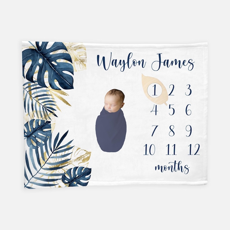 May include: A white baby blanket featuring a blue and gold tropical leaf design. The blanket includes a space for a photo and a numbered chart to track a baby's growth from 1 to 12 months. The text "Waylon James" is printed on the blanket.
