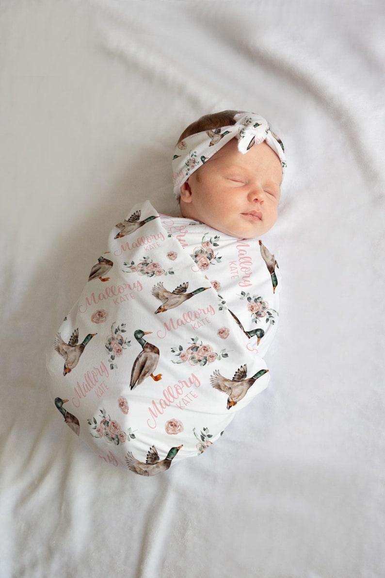 May include: A newborn baby wrapped in a white swaddle blanket and matching headband. The blanket and headband feature a pattern of watercolor ducks, pink roses, and the name "Mallory Kate" in pink script. The baby is lying on a white sheet.