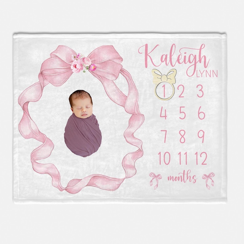 May include: A white baby blanket with a pink bow and floral design. The blanket has a space for a photo and a number chart for tracking a baby's growth. The text "Kaleigh Lynn" is printed on the blanket.