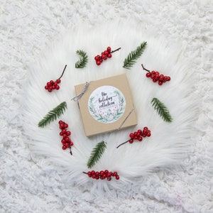May include: A square, light brown box with a silver ribbon, adorned with a circular sticker reading "The Holiday Collection." The box is surrounded by sprigs of red berries and green pine needles, all set on a white, fluffy surface.