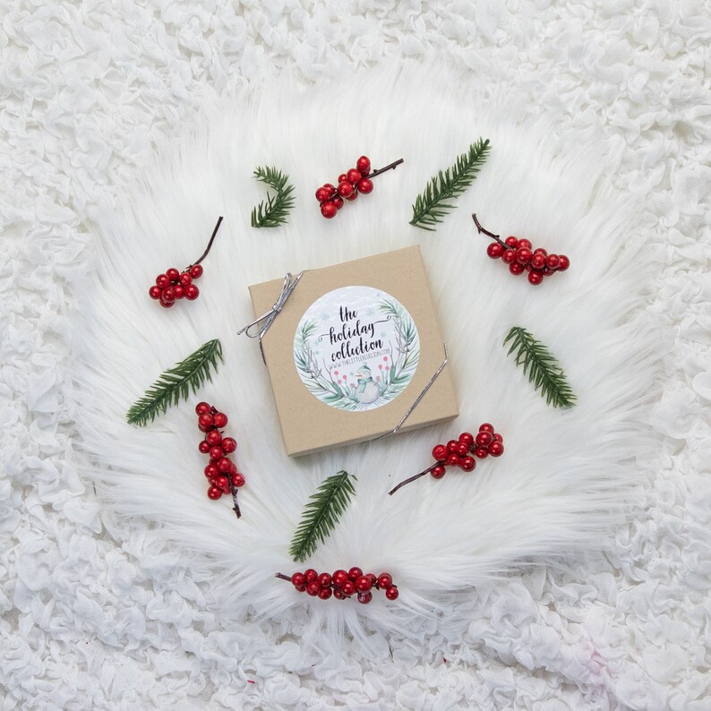 May include: A square, light brown box with a silver ribbon, adorned with a circular sticker reading "The Holiday Collection." The box is surrounded by sprigs of red berries and green pine needles, all set on a white, fluffy surface.