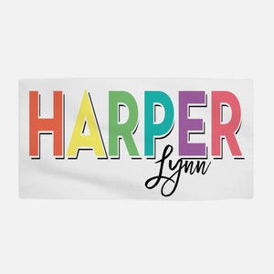 Op de afbeelding: Een witte handdoek met de naam "HARPER" in regenboogkleuren en de naam "Lynn" in zwarte cursieve letters.