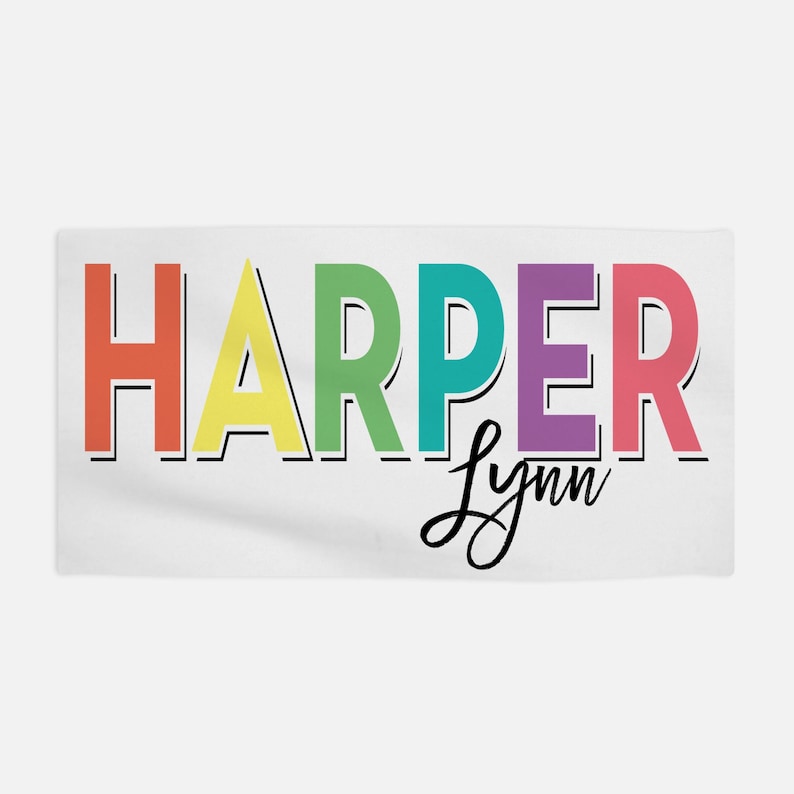 Op de afbeelding: Een witte handdoek met de naam "HARPER" in regenboogkleuren en de naam "Lynn" in zwarte cursieve letters.