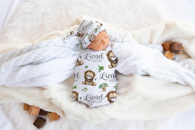 K&ouml;nnte beinhalten: Ein wei&szlig;er Swaddle mit einem L&ouml;wenmuster und dem Namen "Lionel Wilson", der sich wiederholt. Ein Baby ist in die Decke gewickelt und tr&auml;gt eine passende L&ouml;wenm&uuml;tze.