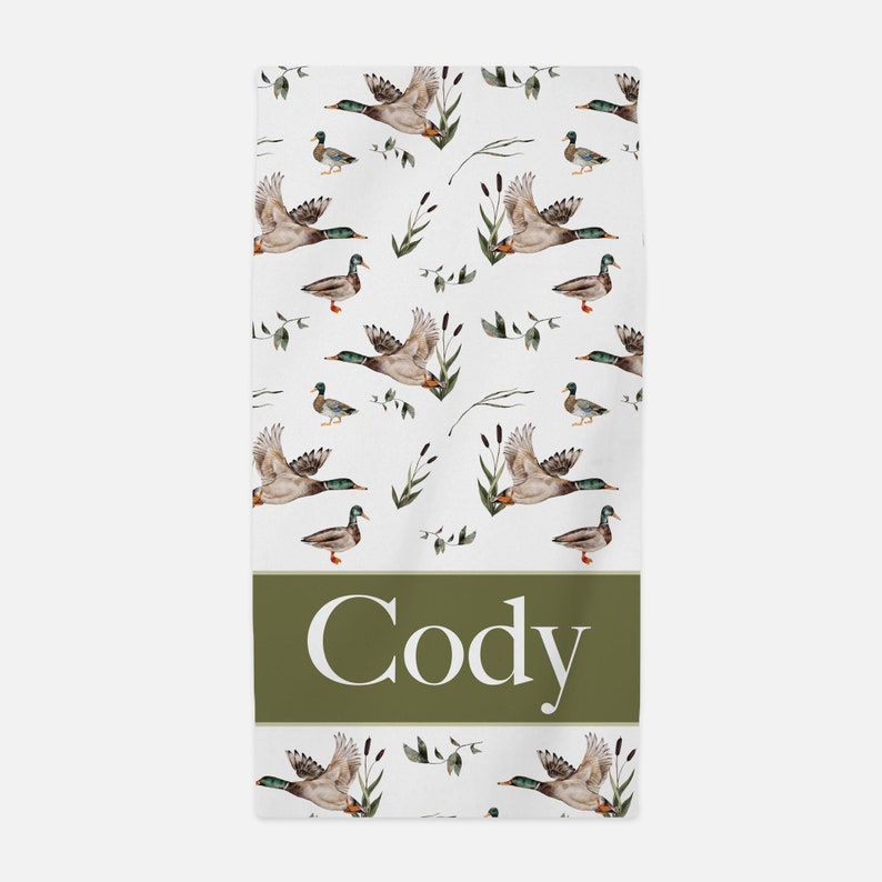 May include: White bath towel with a green border and a pattern of flying ducks and green reeds. The name "Cody" is printed in white on the green border.