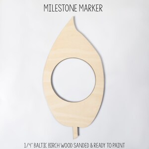 May include: A wooden leaf-shaped milestone marker with a circular cutout. The text "MILESTONE MARKER" is at the top, and "1/4" BALTIC BIRCH WOOD SANDED & READY TO PAINT" is at the bottom. The marker is made of light-colored wood.