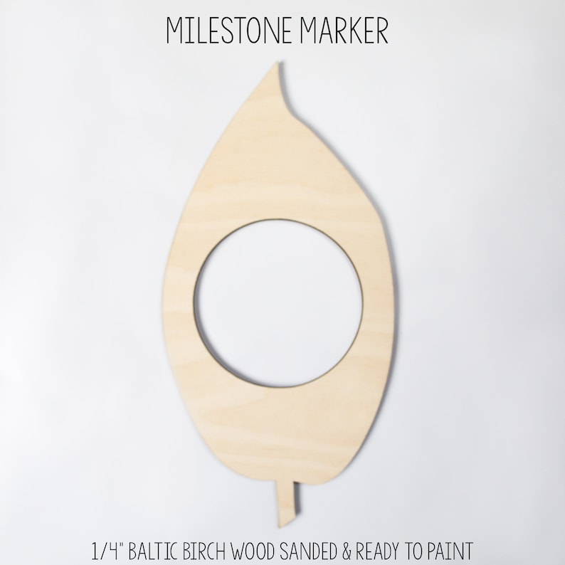 May include: A wooden leaf-shaped milestone marker with a circular cutout. The text "MILESTONE MARKER" is at the top, and "1/4" BALTIC BIRCH WOOD SANDED & READY TO PAINT" is at the bottom. The marker is made of light-colored wood.