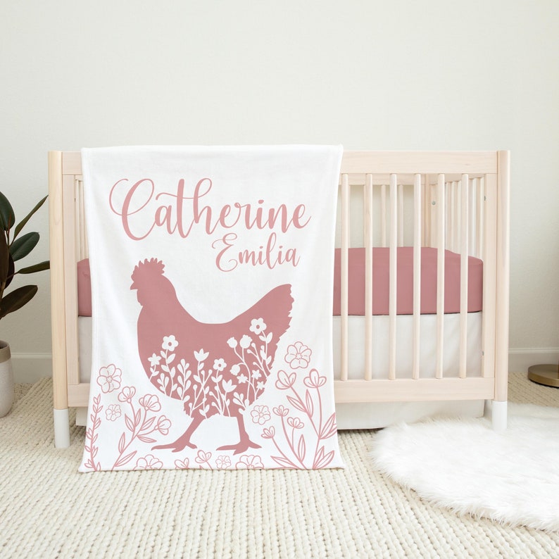 Op de afbeelding: Een witte babydeken met een roze kippensilhouet en een bloemenpatroon. De deken heeft de namen "Catherine Emilia" in roze cursief.