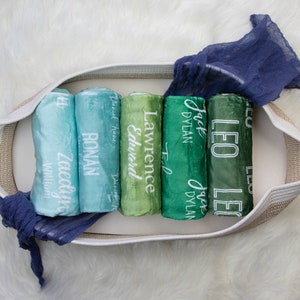 May include: A white wicker basket filled with rolled up green and blue fabric swaddles. Each swaddle is personalized with a name in white lettering. The names are: Zaedyn, Ronan, Lawrence, Edward, Jack Dylan, and Leo.