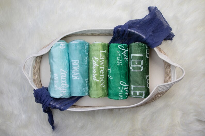 May include: A white wicker basket filled with rolled up green and blue fabric swaddles. Each swaddle is personalized with a name in white lettering. The names are: Zaedyn, Ronan, Lawrence, Edward, Jack Dylan, and Leo.