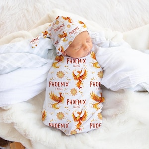 May include: A white baby swaddle and matching hat with a repeating pattern of a phoenix, suns, and the name "PHOENIX LAINE" in orange and yellow. The baby is wrapped in the swaddle and wearing the hat.