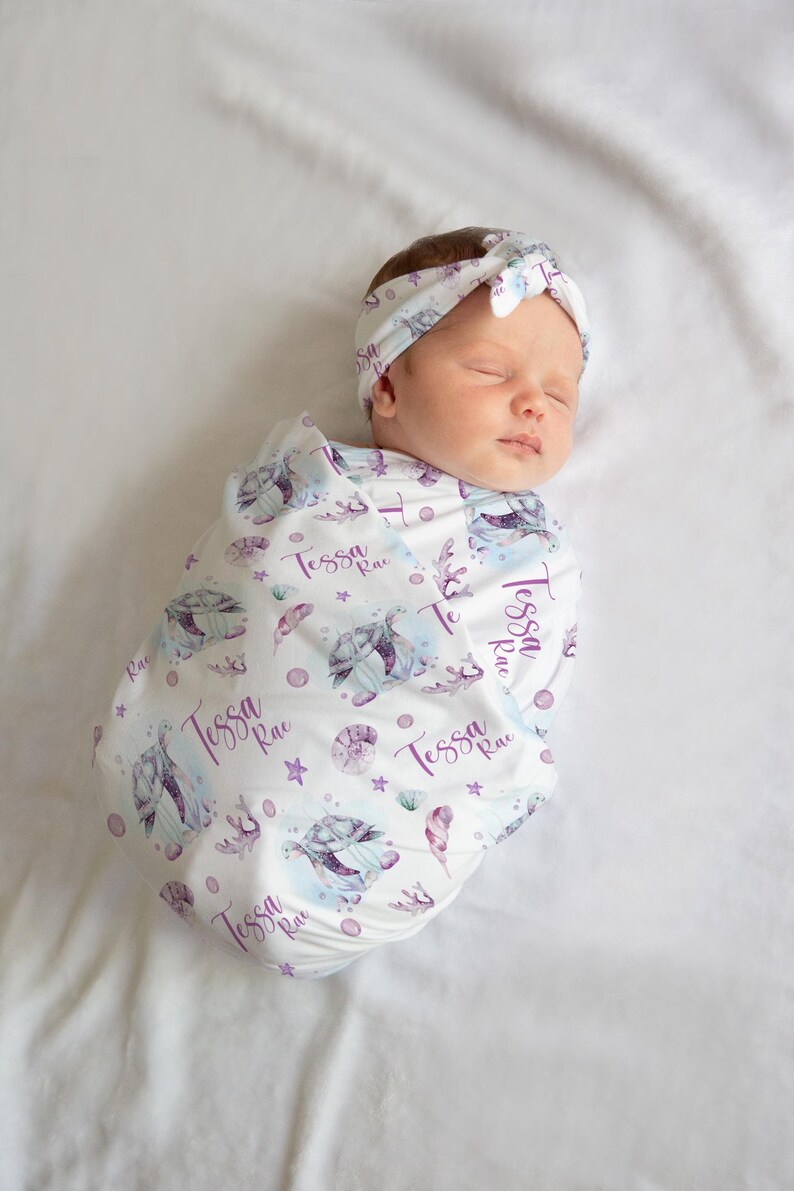 May include: A white swaddle blanket with a purple and blue sea turtle design. The blanket has the name "Tessa Rae" repeated throughout the design. A baby is wrapped in the blanket and wearing a purple headband.