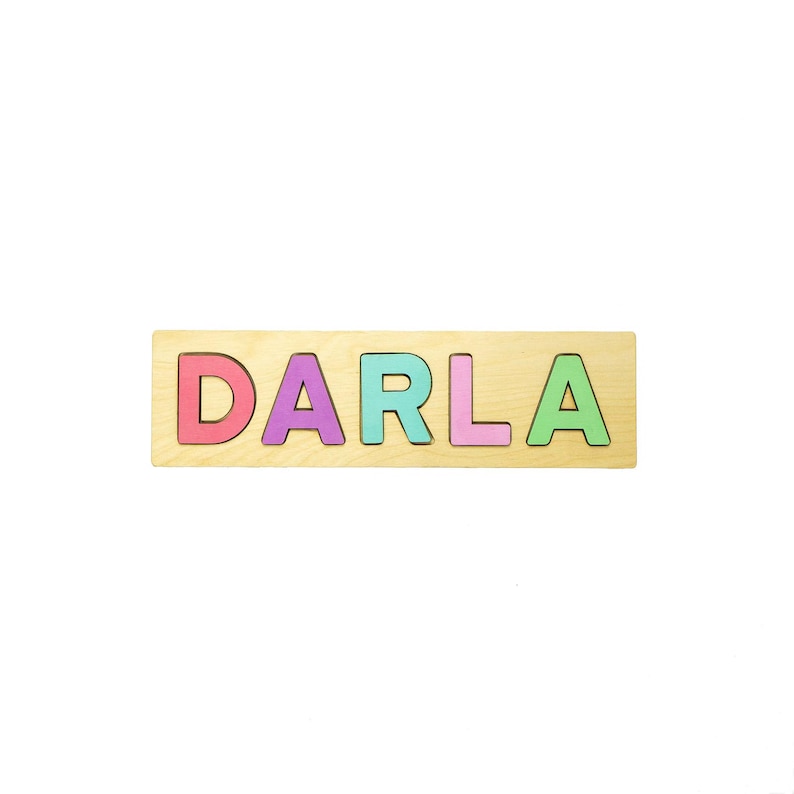 May include: Wooden puzzle spelling the name "DARLA" in colorful, individual letters. The letters are pink, purple, teal, light pink, and green, set against a light wood background. A fun and educational toy for children.