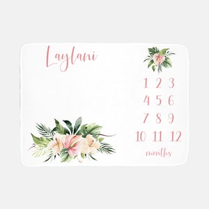 May include: White baby milestone blanket with pink floral accents and the name "Laylani" in cursive. Features numbers 1-12 and the word "months" in pink, with floral arrangements in the corners. Perfect for capturing baby's growth.
