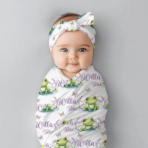 May include: A white swaddle blanket with a green frog and purple flower pattern. The blanket has the name "Willa Rae" repeated on it. A baby is wrapped in the blanket and is wearing a white headband with a purple bow.