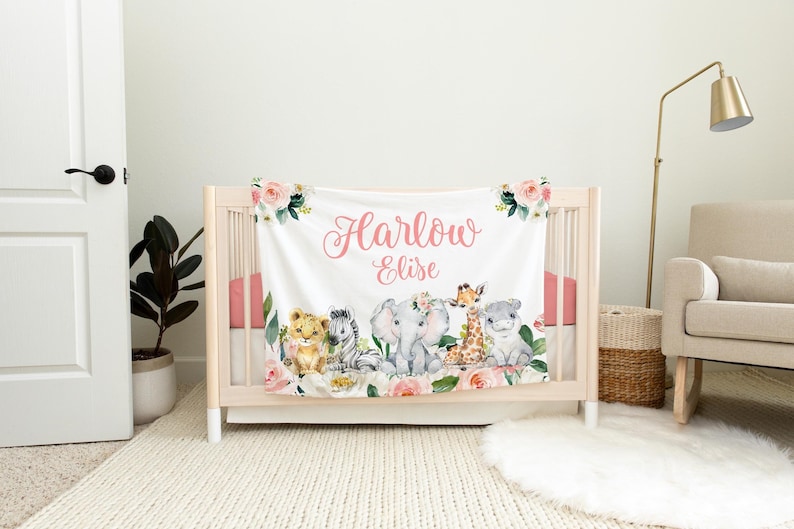 K&ouml;nnte beinhalten: Eine wei&szlig;e Babydecke mit einem rosafarbenen Blumendesign und einem rosafarbenen Namen "Harlow Elise" in Kursivschrift. Die Decke zeigt eine Reihe von niedlichen Tierillustrationen, darunter ein L&ouml;we, ein Zebra, ein Elefant, eine Giraffe und ein Flusspferd.