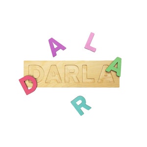 May include: A wooden name puzzle spelling "DARLA" with colorful letters. The letters are pink, purple, green, and teal. The puzzle is made of light-colored wood and is a fun educational toy.