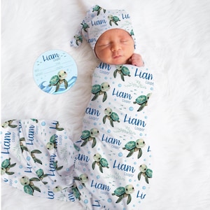 May include: A newborn baby swaddled in a white and blue turtle-themed blanket and matching hat. The blanket and hat are patterned with sea turtles, the name "Liam", and the name "Cooper". A round blue card with a turtle illustration and the name "Liam" is also present.