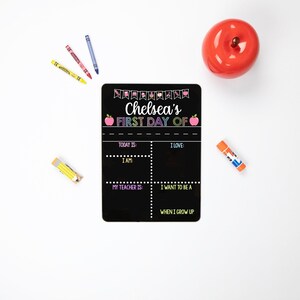 May include: Black chalkboard with a colorful "Chelsea's First Day Of" sign with apple graphics. The chalkboard has sections for writing "Today is", "I am", "My teacher is", "I love", "I want to be a", and "When I grow up".