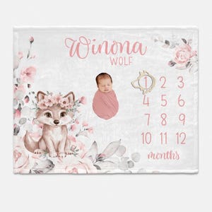 May include: A white baby blanket with a pink floral design and a cute wolf illustration. The blanket has a space for a baby photo and a monthly growth chart with numbers 1-12. The text "Winona Wolf" is printed on the blanket.