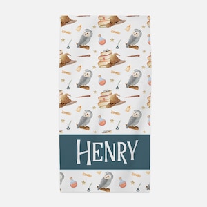 May include: A white bath towel with a blue stripe across the bottom. The towel has a repeating pattern of owls, books, hats, candles, and stars. The name "HENRY" is printed in white on the blue stripe.