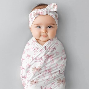 May include: A white swaddle blanket with a pink bow pattern and the name "Kaleigh Lynn" repeated throughout. A baby is wrapped in the blanket and wearing a pink headband with a bow.