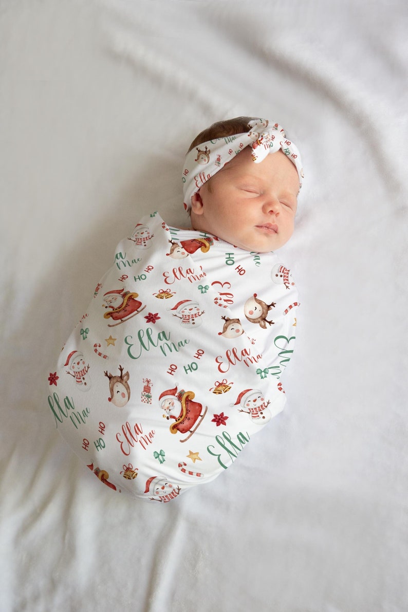 K&ouml;nnte beinhalten: Eine wei&szlig;e Wickeldecke mit Weihnachtsmotiven wie Weihnachtsmann, Rentier, Schneemann und Zuckerstangen. Die Decke tr&auml;gt den Namen "Ella Mae", der sich &uuml;ber das gesamte Design wiederholt. Ein Baby ist in die Decke gewickelt und tr&auml;gt ein passendes Stirnband.