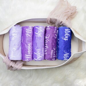 May include: A white wicker basket filled with six rolled up purple and blue blankets with white lettering. The blankets are personalized with names including "Abby", "Kaelyn Marie", "Skyle", "Elliana", "Ellie", and "Sarah Kay".