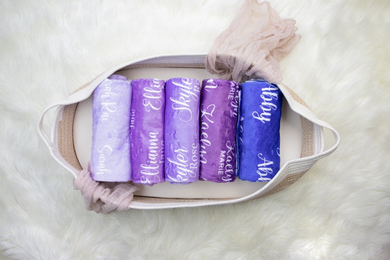 May include: A white wicker basket filled with six rolled up purple and blue blankets with white lettering. The blankets are personalized with names including "Abby", "Kaelyn Marie", "Skyle", "Elliana", "Ellie", and "Sarah Kay".