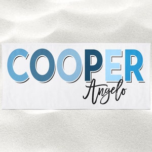 Op de afbeelding: Een witte strandhanddoek met de naam "COOPER" in blauwe letters met een blauw en wit ombre-effect. De naam "Angelo" staat eronder in cursief.