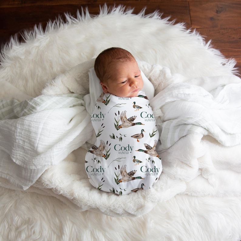 May include: A white swaddle blanket with a green and brown duck print. The blanket has the name "Cody Hunter" repeated on it. A baby is swaddled in the blanket.
