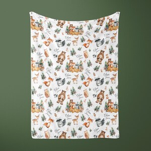 May include: White baby blanket with a woodland animal print. The print features bears, raccoons, deer, owls, and foxes. The blanket has the text "Colin James" repeated throughout the pattern.