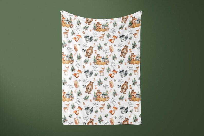 May include: White baby blanket with a woodland animal print. The print features bears, raccoons, deer, owls, and foxes. The blanket has the text "Colin James" repeated throughout the pattern.