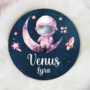 May include: A round, navy blue decorative piece featuring a watercolor illustration of an astronaut on a crescent moon. Pink stars and a rocket surround the astronaut. The text "Venus Lyra" is written in white script.