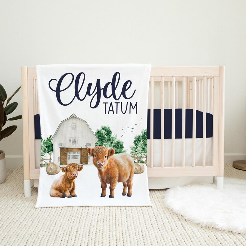 Op de afbeelding: Een witte babydeken met een rode schuur, groene bomen en twee bruine Highland-koeien. De deken heeft de naam "Clyde Tatum" in zwarte cursieve letters.