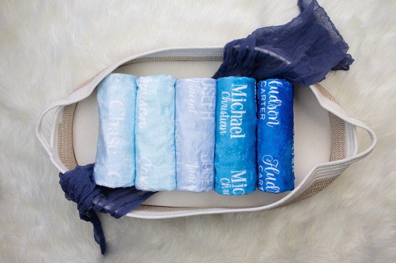 May include: A set of six blue and white personalized baby blankets with names embroidered on them, arranged in a woven basket.