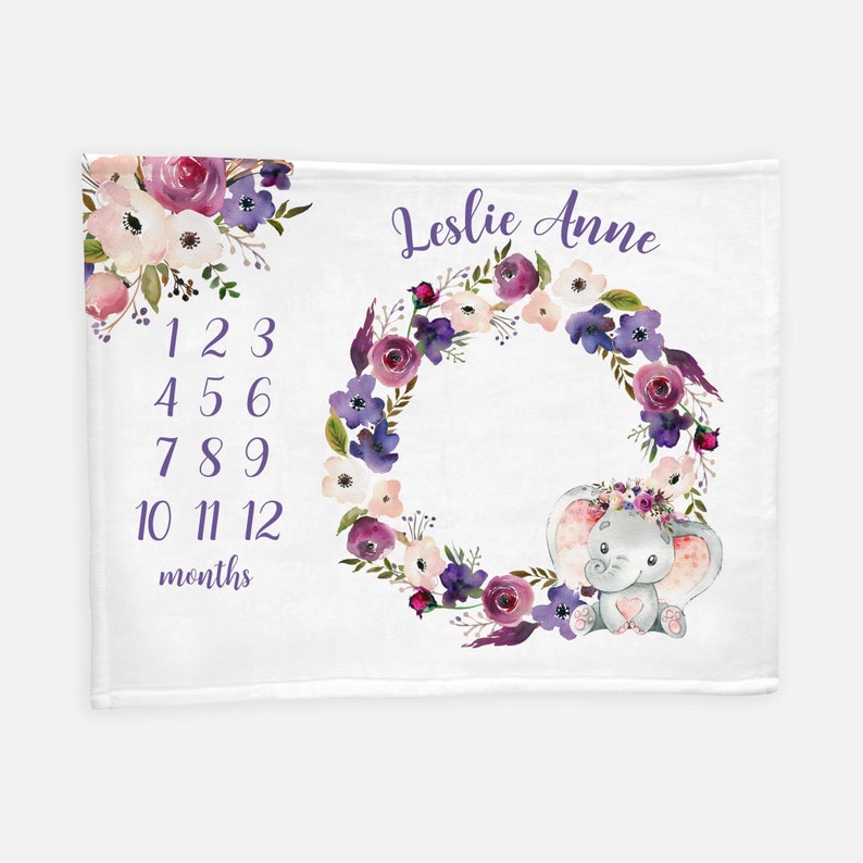 May include: A white baby blanket with a purple floral wreath and a gray elephant with a pink heart. The blanket has a number chart for tracking a baby's growth and the name "Leslie Anne" is written at the top.