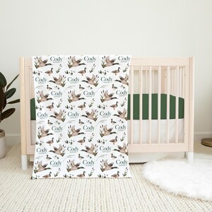 May include: A white baby blanket with a repeating pattern of ducks and the name "Cody Hunter" printed on each duck. The blanket is in front of a wooden crib with a green mattress.