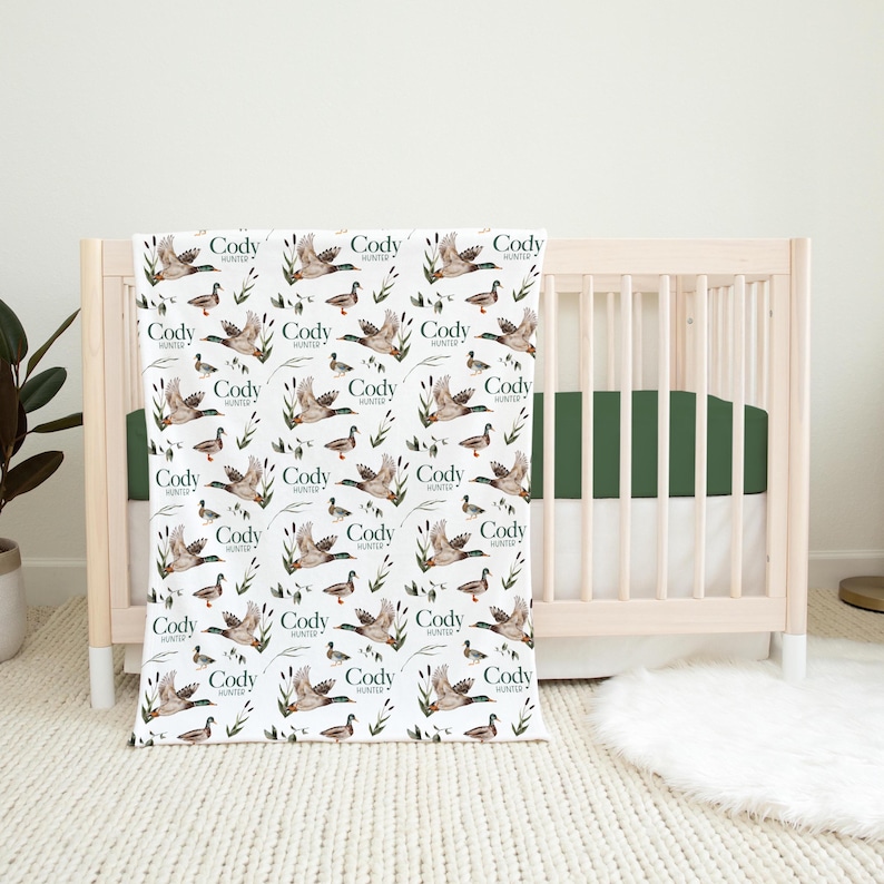 May include: A white baby blanket with a repeating pattern of ducks and the name "Cody Hunter" printed on each duck. The blanket is in front of a wooden crib with a green mattress.