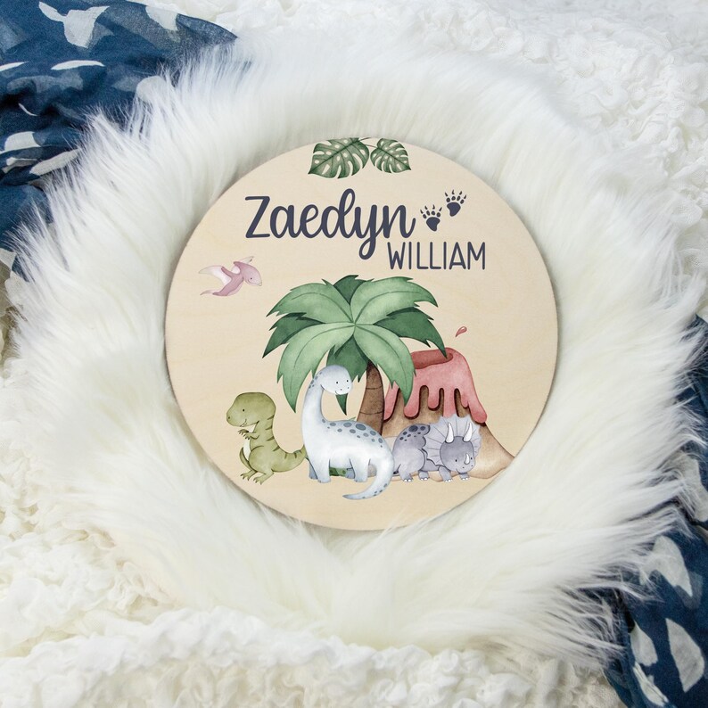 K&ouml;nnte beinhalten: Ein rundes Holzschild mit Dinosaurier-Motiv. Das Schild zeigt einen gr&uuml;nen Palmenbaum, einen Vulkan und vier Dinosaurier. Der Text "Zaedyn William" ist in Lila auf dem Schild geschrieben.