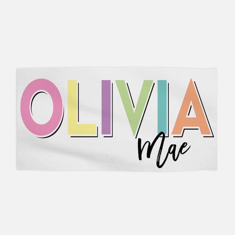 Op de afbeelding: Een witte handdoek met de naam "Olivia Mae" geschreven in kleurrijke blokletters. De letters zijn zwart omlijnd en gevuld met verschillende pastelkleuren.
