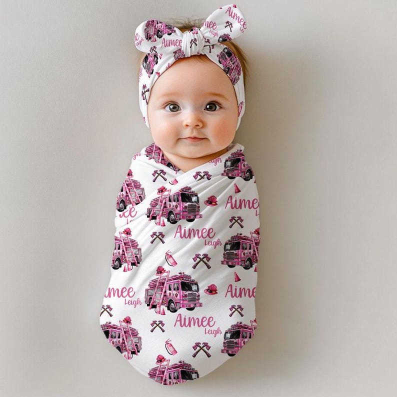 Pu&ograve; includere: Una copertina bianca per neonati con un motivo di camion dei pompieri rosa. La copertina riporta il nome "Aimee Leigh" ripetuto su tutto il motivo. Un cerchietto rosa con fiocco &egrave; sulla testa del bambino.