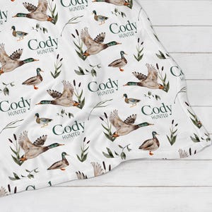 May include: White baby blanket with a repeating pattern of green leaves, brown ducks, and the name "Cody Hunter" in a dark green font.