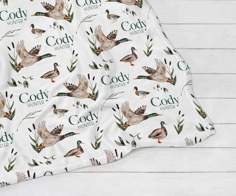 May include: White baby blanket with a repeating pattern of green leaves, brown ducks, and the name "Cody Hunter" in a dark green font.